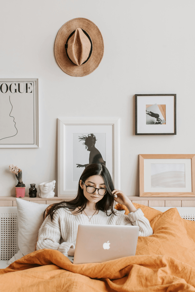 A woman sitting on her bed with a n orange blanket, typing on a laptop
