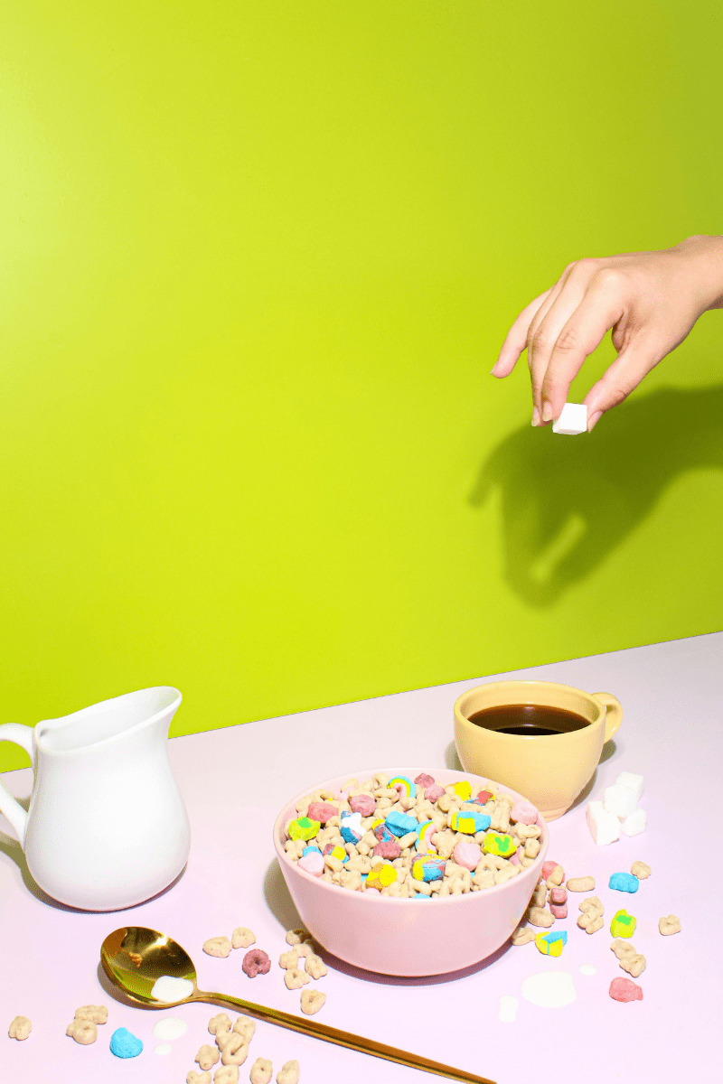 Vibrant green background covered in cereal, a milk pitcher, and a cup of coffee. A hand is dropping sugar cubes into the coffee, next to a spoon